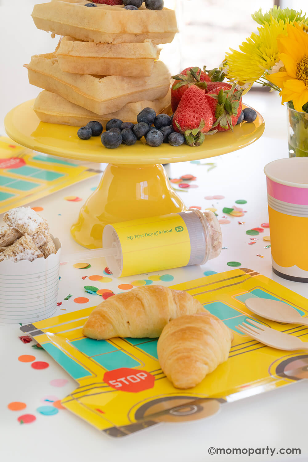 A first day of school breakfast celebration table with delicious breakfast food including Belgium waffles stacked as a cake with mixed berries, on the table is a school bus shaped party plates with mini croissants for kids, along with pencil shaped party cups and pencil party poppers, these back to school party ideas by Momopartys make a great inspiration for any school themed celebrations with your kids at home.