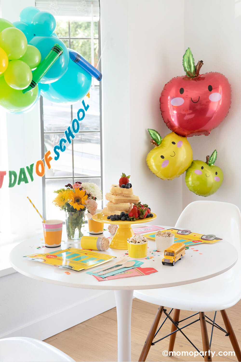 A kitchen table area decorated with back to school themed balloons in fun, bright colors and an apple stacker foil balloon in a adorable illustration appealing to kids. On the table there is a stack of Belgium waffles as the "breakfast cake" and multiple school themed tableware from momopartys.shop. All in all creates a great look and inspiration for kid's back to school celebration or party.