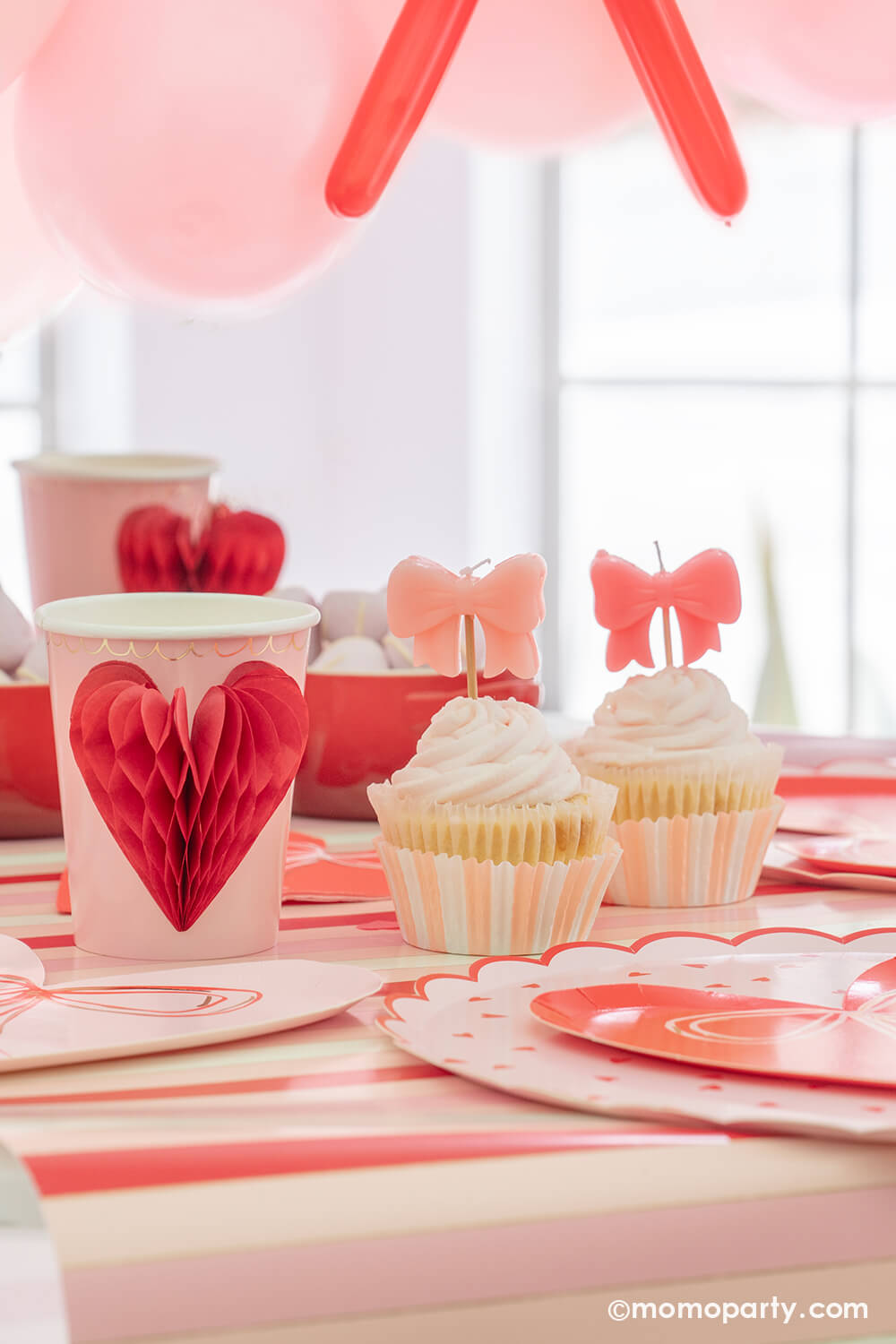 A close-up detail of Momopartys's sweet Pink and Red Bow-Themed Valentine's Day Party table. Highlighting Meri Meri Honeycomb Heart Cups, these cups feature charming 3D honeycomb red hearts on pink paper, adorned with gold foil details. Accompanied by a Pink Bow Candles atop a cupcakes and sweets in the red Mini Cocotte, with a flower vase behind them. All elegantly arranged on a red-striped paper runner. Such a cute idea for elevating your Valentine's celebration or Galentine's Day with your besties.
