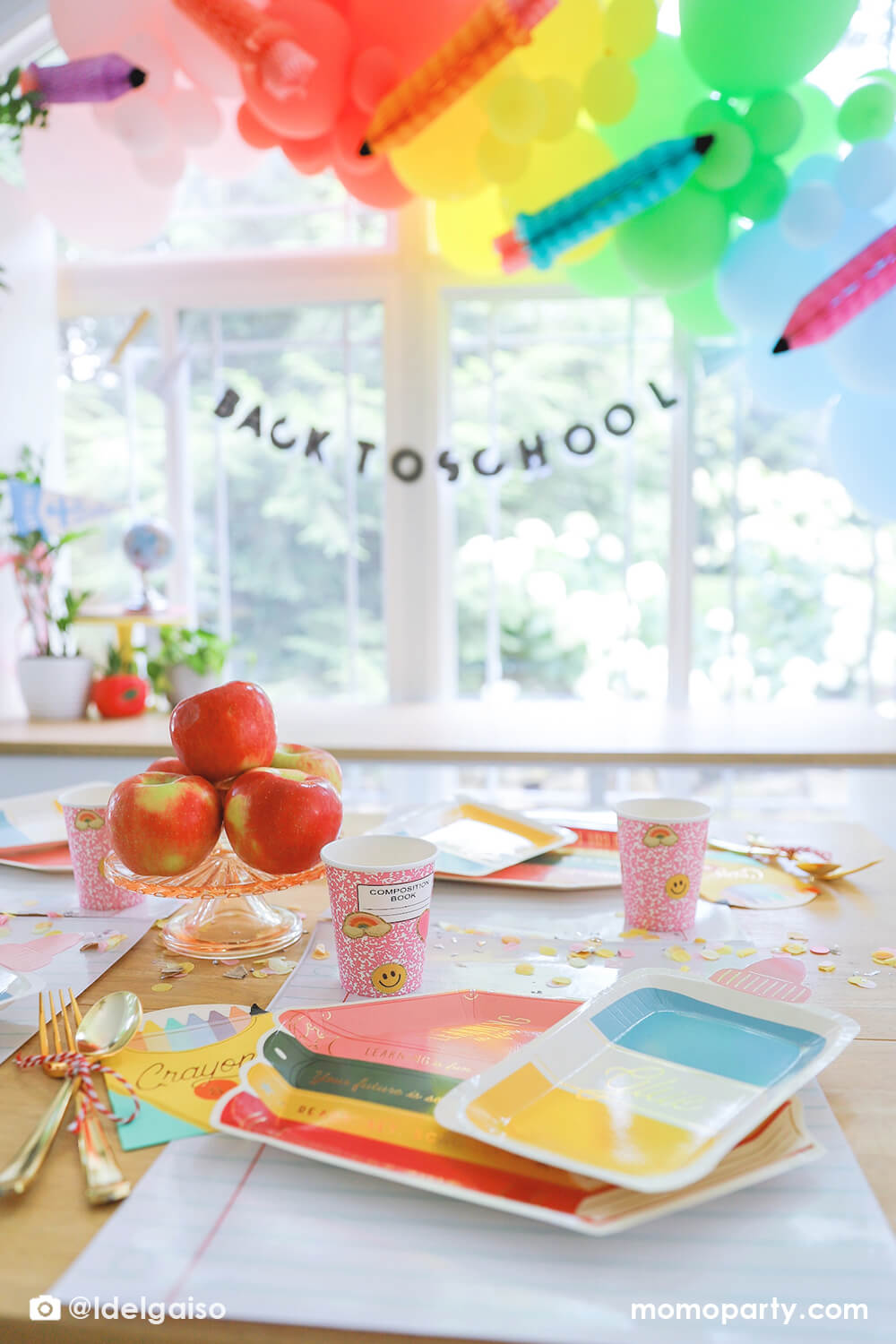 A festive back to school party featuring adorable school themed party supplies from Momopartys including the colorful stack of books shaped plates, glue bottle shaped plates, crayon shaped napkins, pink notebook party cups, giant notebook placemats and pencil inspired confetti around. In the back there's a pastel balloon garland in rainbow colors adorned with matching pencil honeycomb, along with back to school banner hung underneath. Making this a perfect inspo for kid's first day of school celebration.