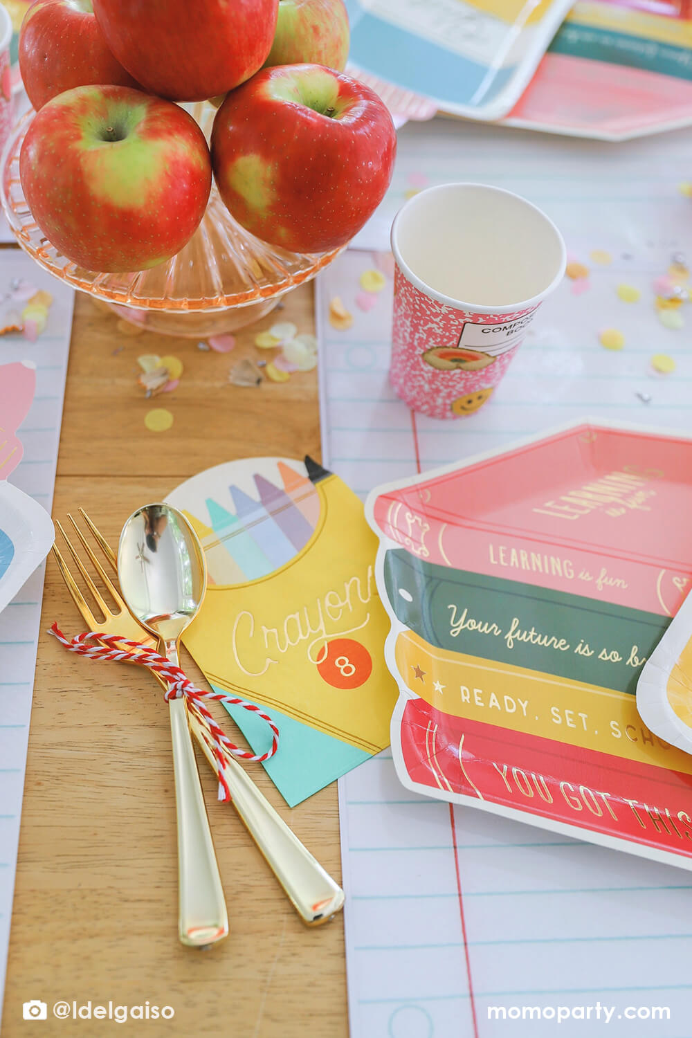 A closeup shot of Momopartys's crayon box shaped napkin and stack of books shaped plate on the giant notebook placemat from a back to school party table. Around plate is a pink notebook inspired party cup and some pencil inspired confetti around it. Along with the stack of apples in the middle of the table as the centerpiece, this simple table setting is perfect for kid's first day of school celebration.