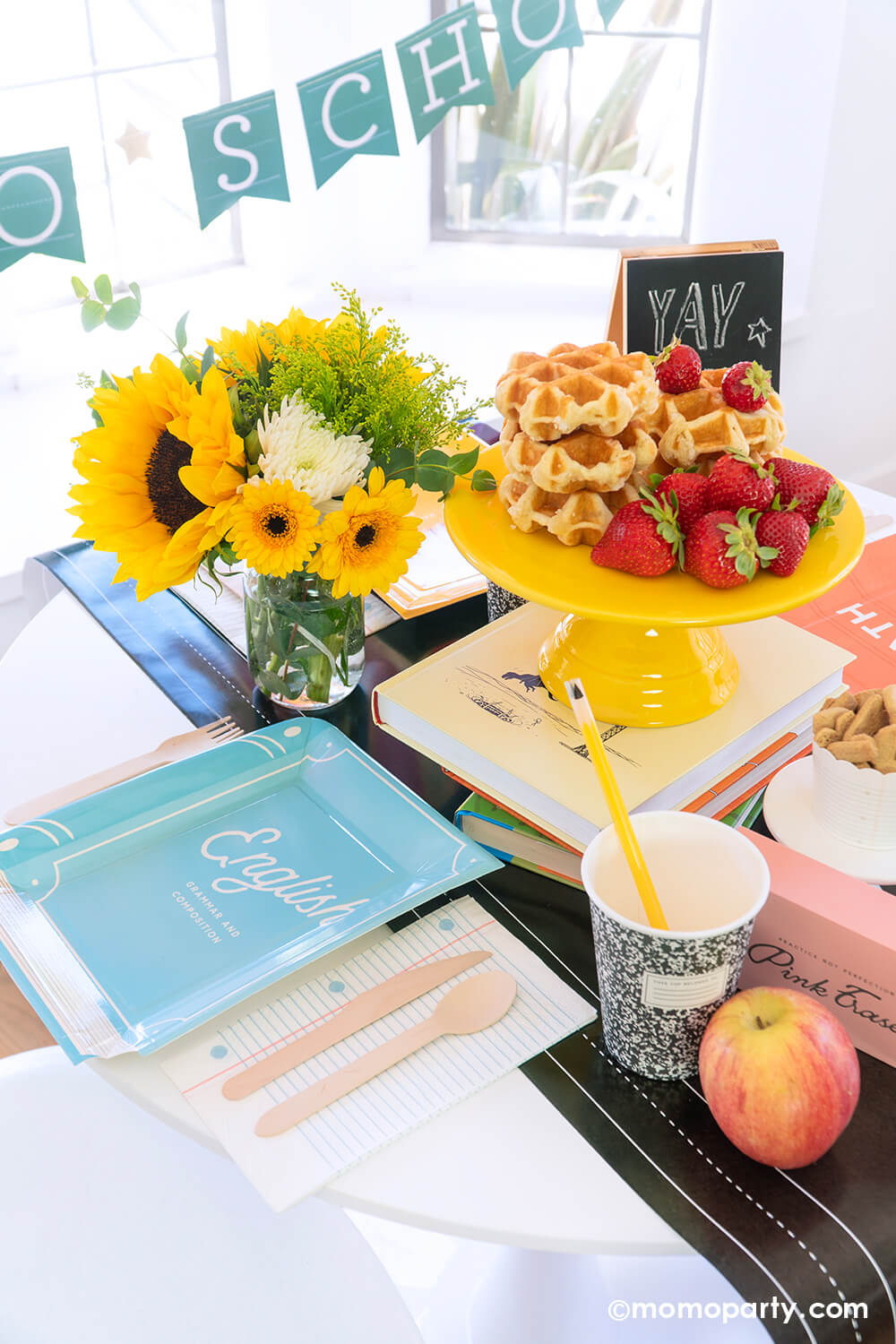 An overhead shot of a back to school party table featuring multiple school themed party goods from Momopartys, including text book shaped plates in different colors, eraser shaped treat boxes, notebook napkins, composition party cups with pencil straws, all on the chalkboard table runner. With stacked waffles on a cake stand, treats, apples, fruits and a sunflower floral arrangement on the table, this makes a perfect idea for your child's back to school party or first day of school celebration!