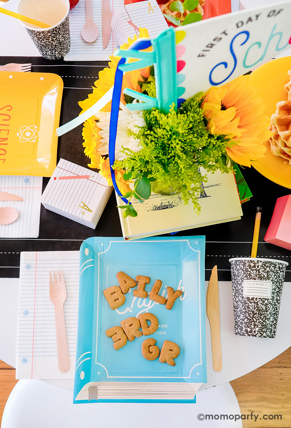 An overhead shot of a first day of school celebration table featuring multiple school themed party goods from Momopartys, including text book shaped plates in different colors, eraser shaped treat boxes, notebook napkins, composition party cups with pencil straws, all on the chalkboard table runner. With stacked waffles, treats, apples, alphabet cookies spelling Baily 3rd Grade on the textbook shaped plate, this makes a perfect idea for your child's back to school party or first day of school celebration!