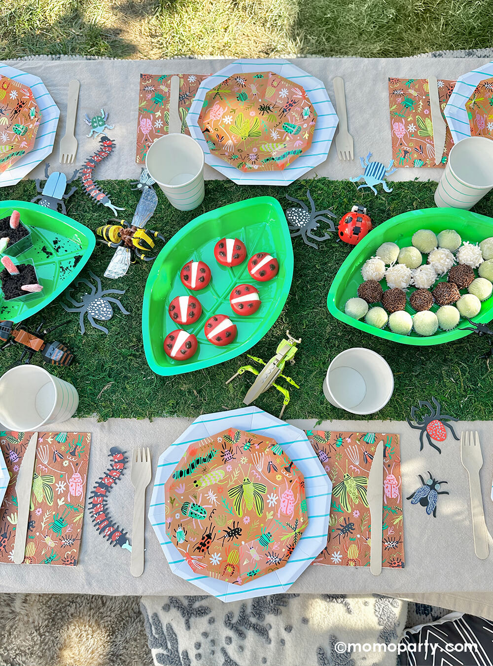 An insect bug themed birthday party table styled by Momopartys features backyard bug plates, napkins with aqua striped plates and party cups by Daydream Society. In the middle there's a faux grass table runner that was decorated with various lego insect toys and palm leaf shaped serving tray filled with insect themed treats and snacks including gummy worms, ladybug babybel cheeses and bug egg shaped brigadeiros, some playful and fun ideas for kid's insect themed bash.