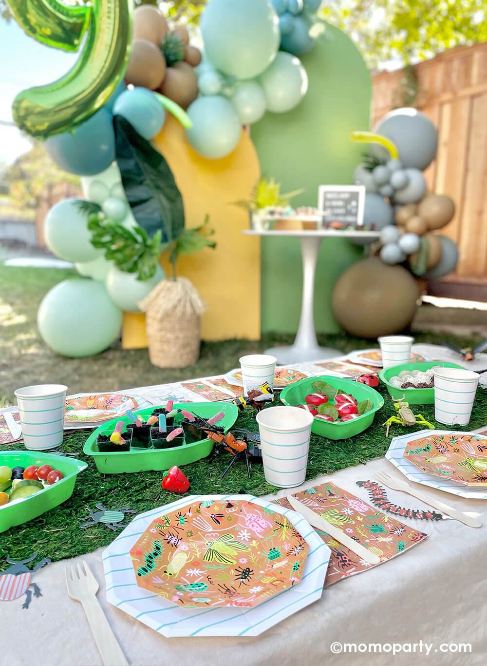An insect bug themed birthday party table styled by Momopartys features backyard bug plates, napkins with aqua striped plates and party cups by Daydream Society. In the middle there's a faux grass table runner that was decorated with various lego insect toys and palm leaf shaped serving tray filled with insect themed treats and snacks including gummy worms, ladybug cheeses, with a balloon backdrop in green, yellow and brown setting a natural tone for this bug themed party.