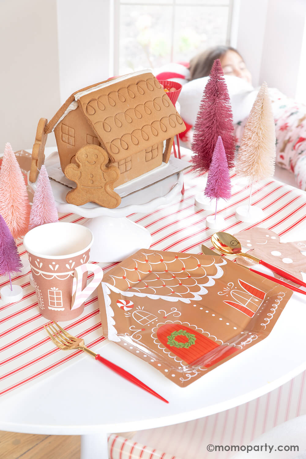A delightful scene at Momopartys's Gingerbread House Decoration Home Party: A dining table adorned with My Mind's Eye Gingerbread House-Shaped Plates, Smiley Gingerbread Man Napkins, Gold and Red Cutlery Set, and Gingerbread House Paper Party Cups with Handles. Also, Gingerbread House cookies are showcased on a cake stand, surrounded by Christmas Bottle Brush Trees, all set atop the festive Believe Christmas Red Striped Table Runner. What a fun idea to celebrate your holiday with kids at home.