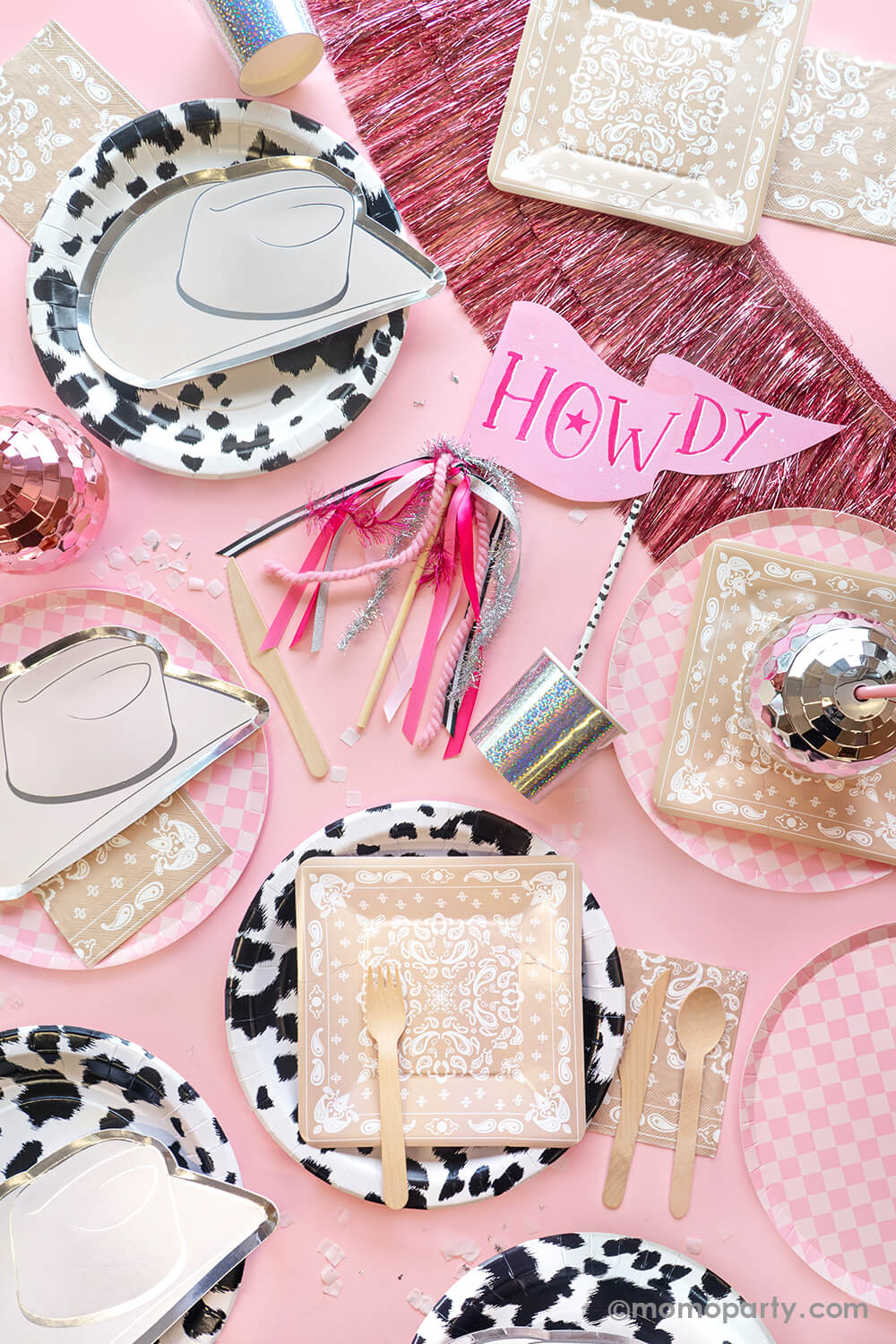 A top view of Momopartys's disco cowgirl party collection! With the howdy party pennant in the middle. surrounding it, featuring silver felt hat dessert plates, layered with black cowhide dinner plates, dusty champagne bandana dessert plates, and check it! pink checkered dinner plates, Also some dusty champagne bandana small napkins, silver and rose gold disco ball tumblers. pink tinsel fringe garland adds a touch of sparkle, making these modern party wares perfect for a disco cowgirl themed party