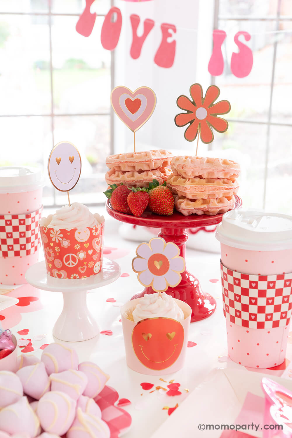 A close up shot of Momopartys's Groovy Valentine's party box featuring My Mind's Eye's love baking cups and toppers set in retro design of daisies, peace signs, smiley faces and hearts. In the back are some heart shaped pink waffles topped with retro toppers with strawberries on the side. Above the table hung "love is all you need" party banner in retro font type, with retro inspired tableware, it makes a perfect inspo for a fun and groovy Valentine's Day celebration!