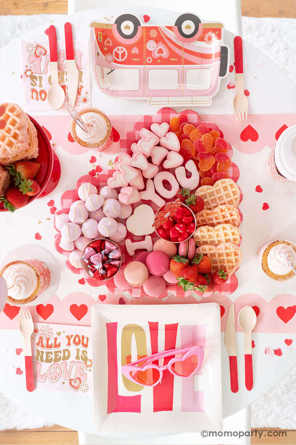 A party table featuring Momopartys's Groovy Valentine's party box with My Mind's Eye's retro Valentine's party supplies including LOVE square plates, VW bus shaped plates, baking cup and topper set and "all you need is love" napkins. On the table there's a heart shaped sweet board filled with Valentine's Day themed treats including "I Love You" sugar cookies, macarons, strawberries, heart shaped marshmallows, heart shaped pink waffles and candies, a perfect inspo for a fun Valentine's celebration!