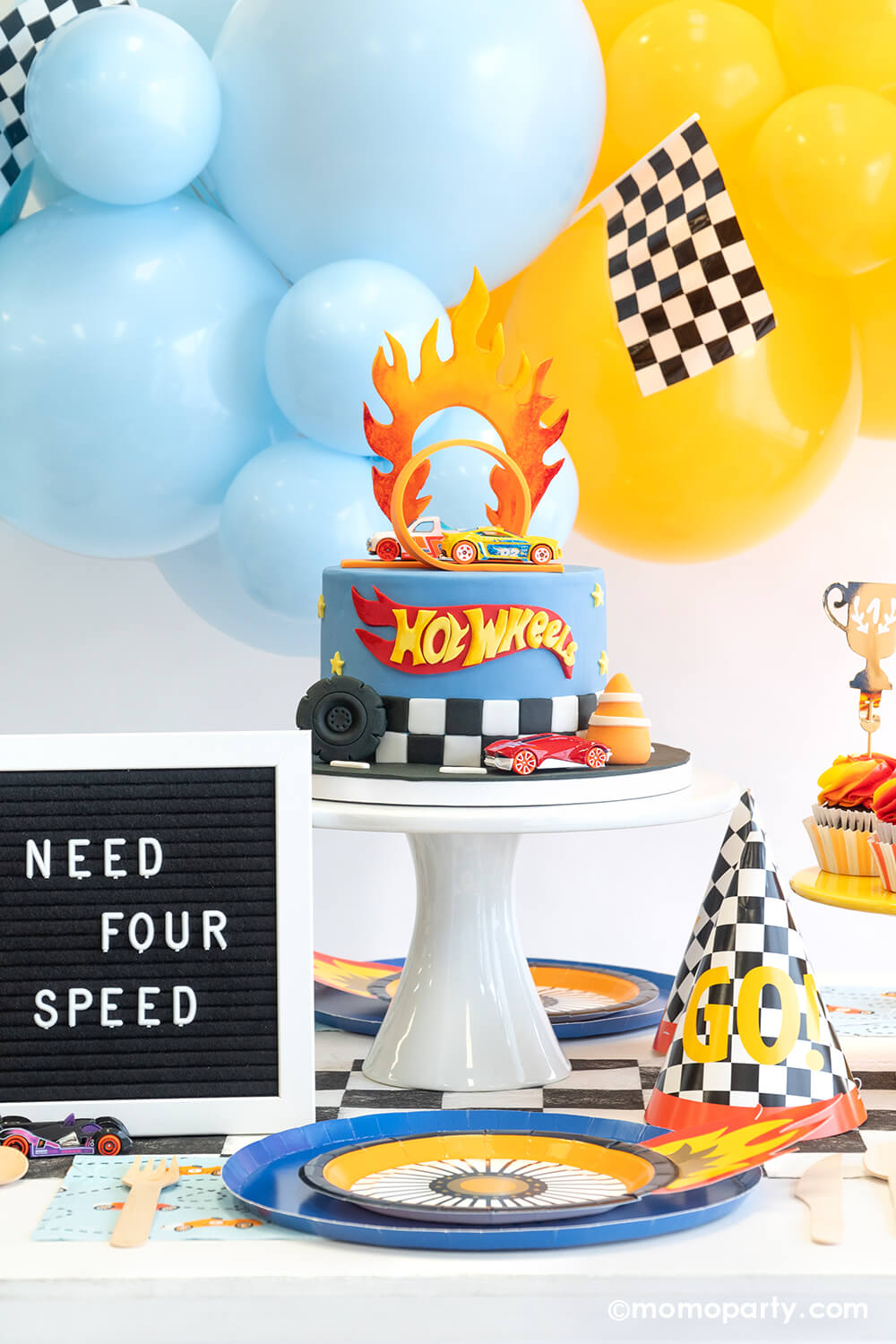 A "Need For Speed" boy's race car themed fourth birthday party featuring Hot Wheels balloon garland, Hot Wheels themed party supplies. A festive party table features Momopartys's Midnight Blue Dinner Plates, Wheel Shaped Plates, checkered party cups and checkered GO! birthday hats, with a Hot Wheels fondant birthday cake.