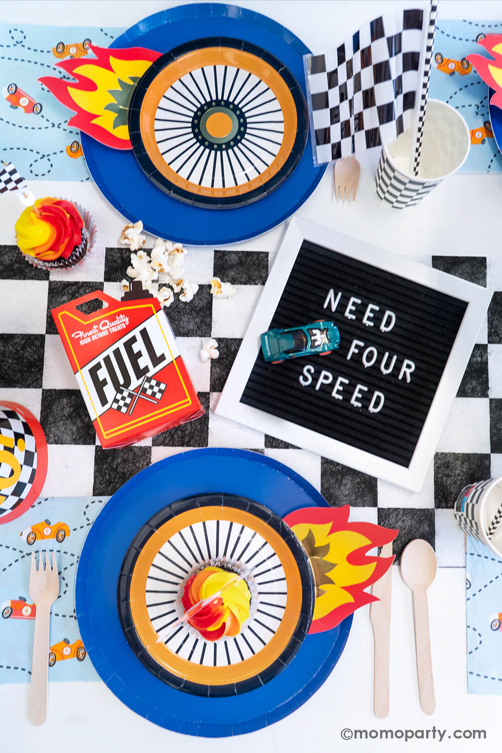 A flatlay shot of a festive "Need FOUR Speed" Hot Wheels themed fourth birthday party table featuring Momopartys's midnight dinner plates with wheel shaped plates, checkered table runner, party cups, paper straws, birthday hats, Fuel gas shaped treat box with popcorn and blue race car napkins . In the middle of the table, there's a black letterboard spelling "NEED FOR SPEED" and a Hot Wheels toy car next to it - a perfect inspo for boy's 4th birthday party.