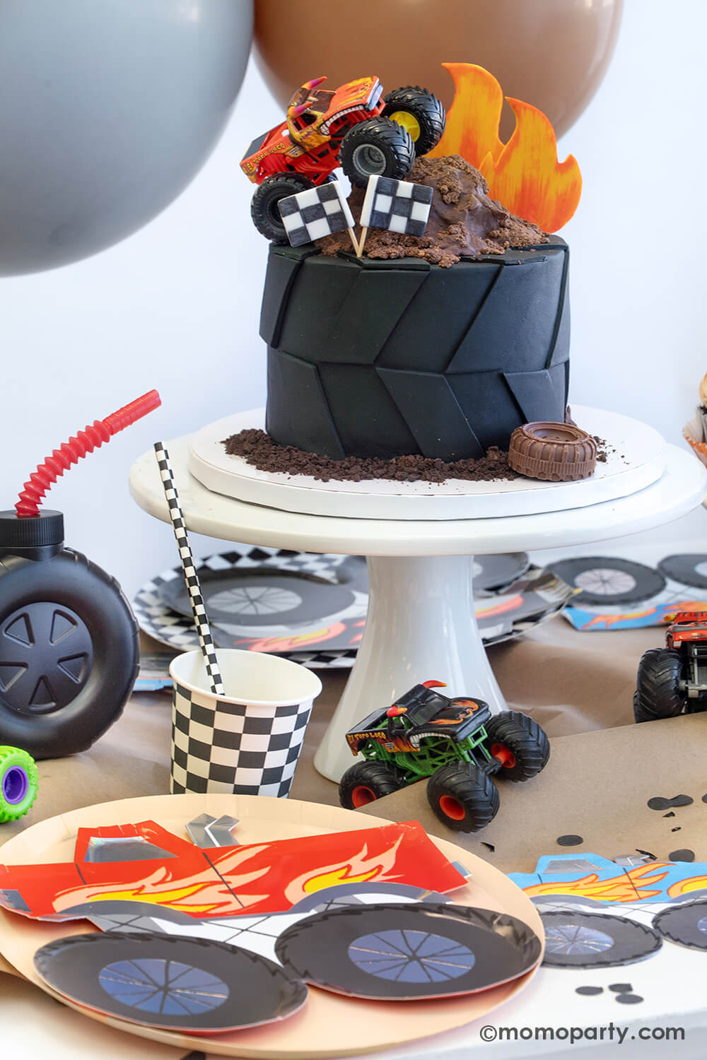 A monster truck themed party table setting styled by Momopartys. The table features a large fondant tire shaped birthday cake topped with a fondant flame, chocolate mud, two race car flags and a chocolate tire candy with some oreo crumbs around it. In The table is covered with brown kraft paper with various monster truck themed tableware including plates, napkins, party cups and straws from Momopartys. With a party balloon garland hung above the table and colorful monster truck toys around.