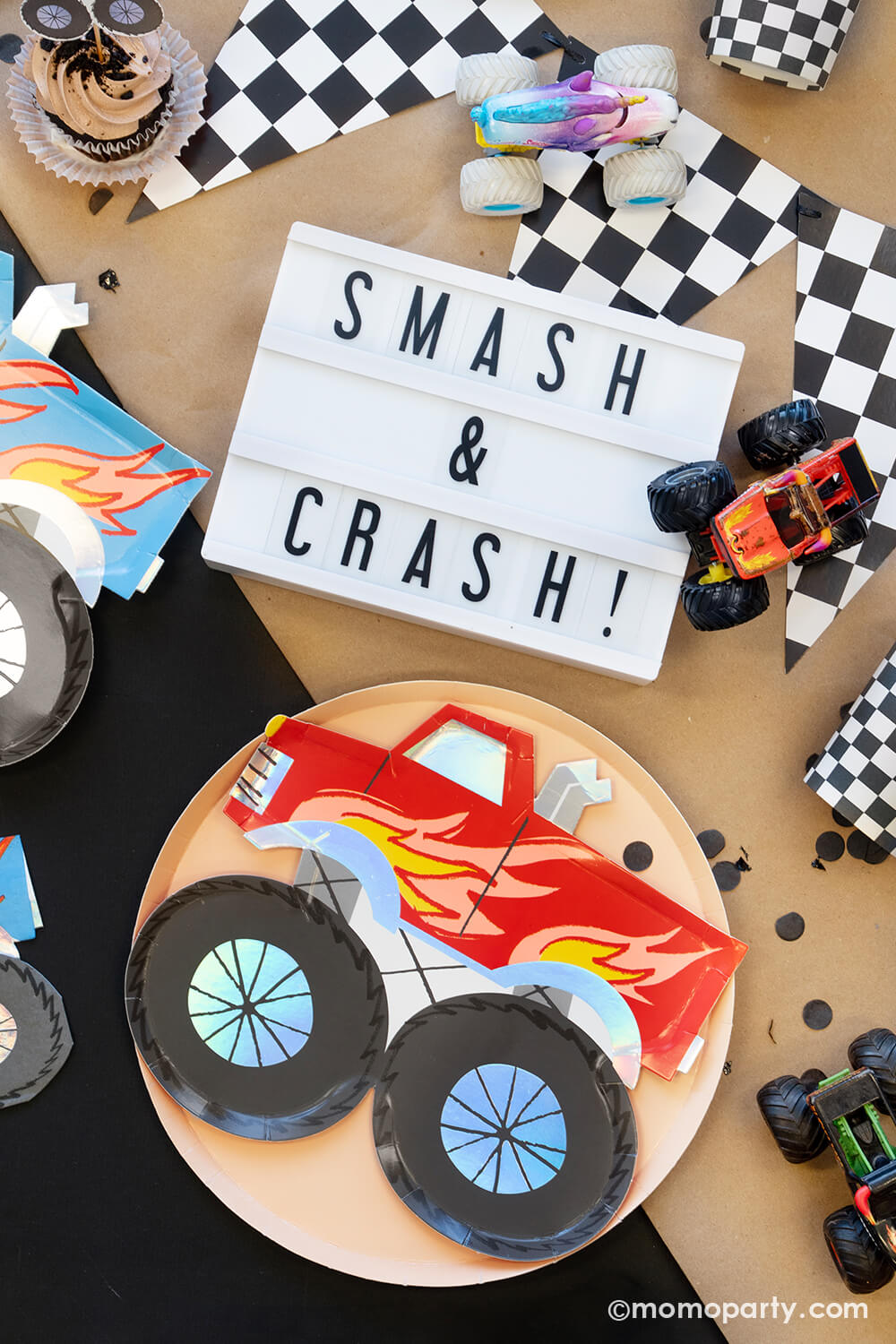 A close up of an overhead shot of Momopartys's Monster Truck Party Collection featuring various monster truck themed tableware and decors including a race car flag banner, black, sand and checkered round dinner plates, party cups, straws and monster truck shaped plates and napkins, with monster truck shaped cupcake kit and "Smash & Crash" sign, monster truck toys on a brown craft paper runner, this collection is perfect for your little monster truck fan's birthday celebration!