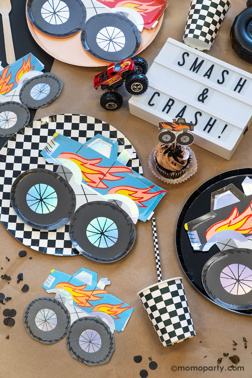 A overhead shot of a monster truck themed party tabescape featuring various monster truck themed tableware from Momopartys including round black and checkered dinner plates, party cups, straws and monster truck shaped plates and napkins, with a brown cupcake topped with a monster truck topper and "Smash & Crash" sign plus truck toys, this setup is perfect for your little monster truck fan's birthday celebration!