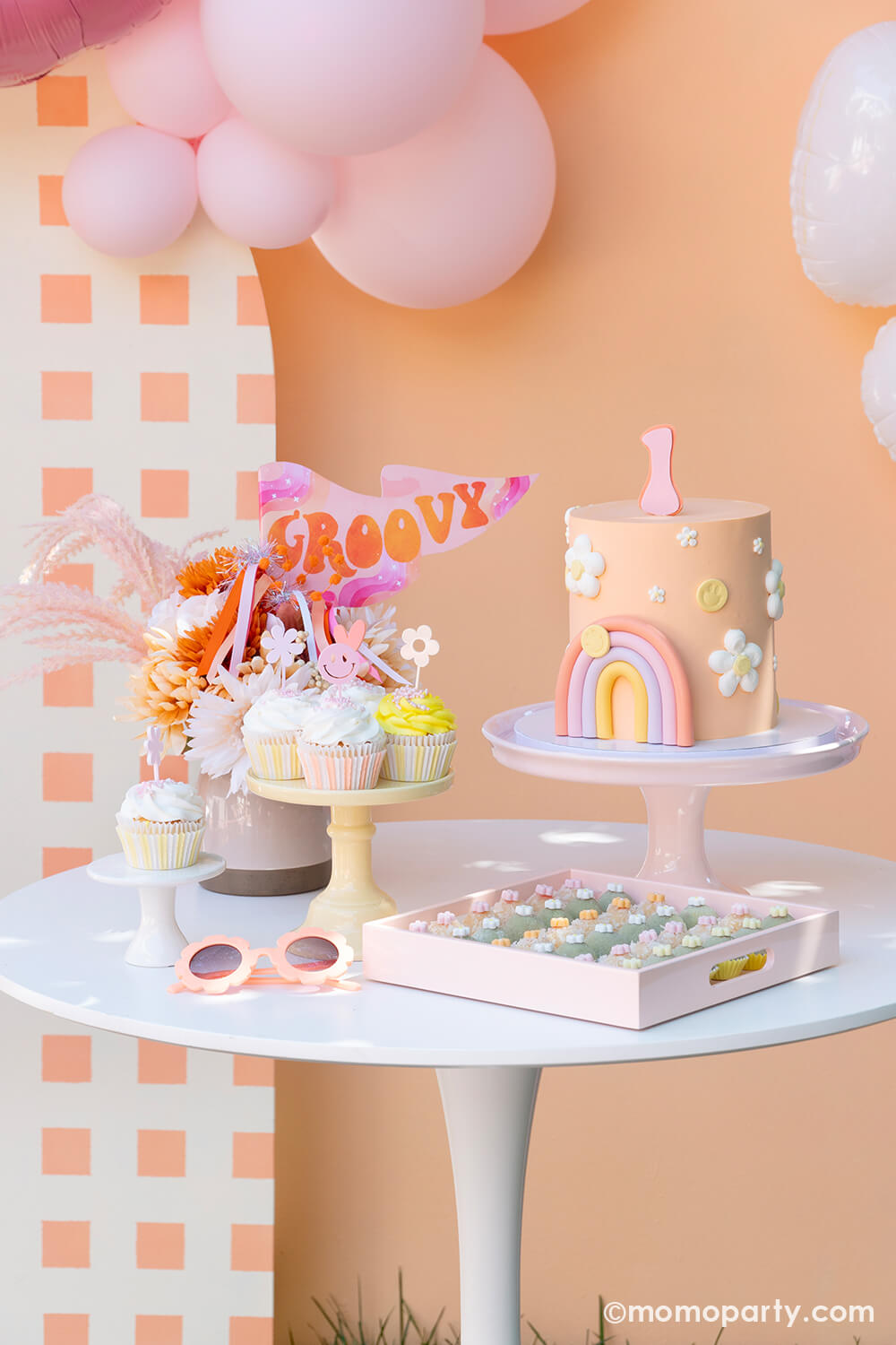 Momo Party’s One Groovy Baby First Birthday Party Dessert Table. Decorated with a modern-themed birthday cake, with pastel rainbow, flower, and smiley face decorations on a peach-colored buttercream cake, a Groovy Party Pennant inside a flower vase, cupcakes with Pastel Retro Acrylic Groovy Cupcake Toppers, peach color sunglasses, a pastel pink tray with Brazilian sweets. Table in front of a check-patterned wooden Backdrop board and balloons. Such a modern and fun for a One Groovy Baby-themed birthday party