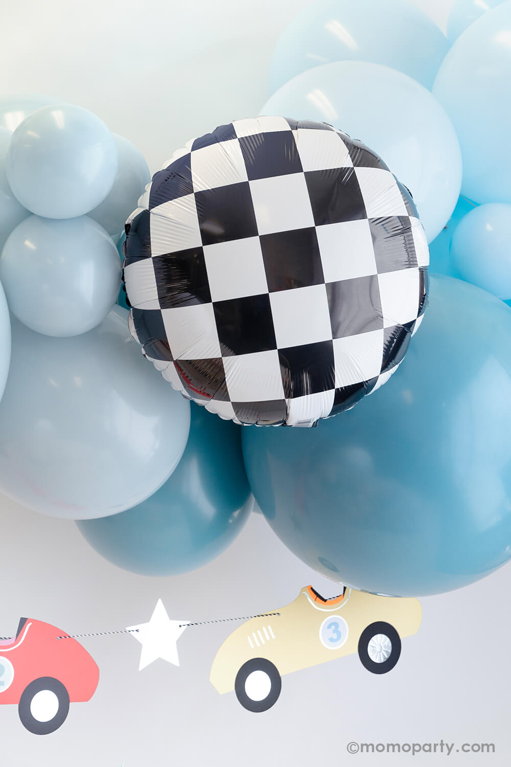 A modern race car themed party decoration by Momopartys featuring a blue balloon garland adorned with checkered foil balloons, underneath hung a vintage race car party garland in pastel colors by Meri Meri. Makes this a perfect party decoration for kid's "Two Fast" second birthday party or a "Fast One" first birthday party.
