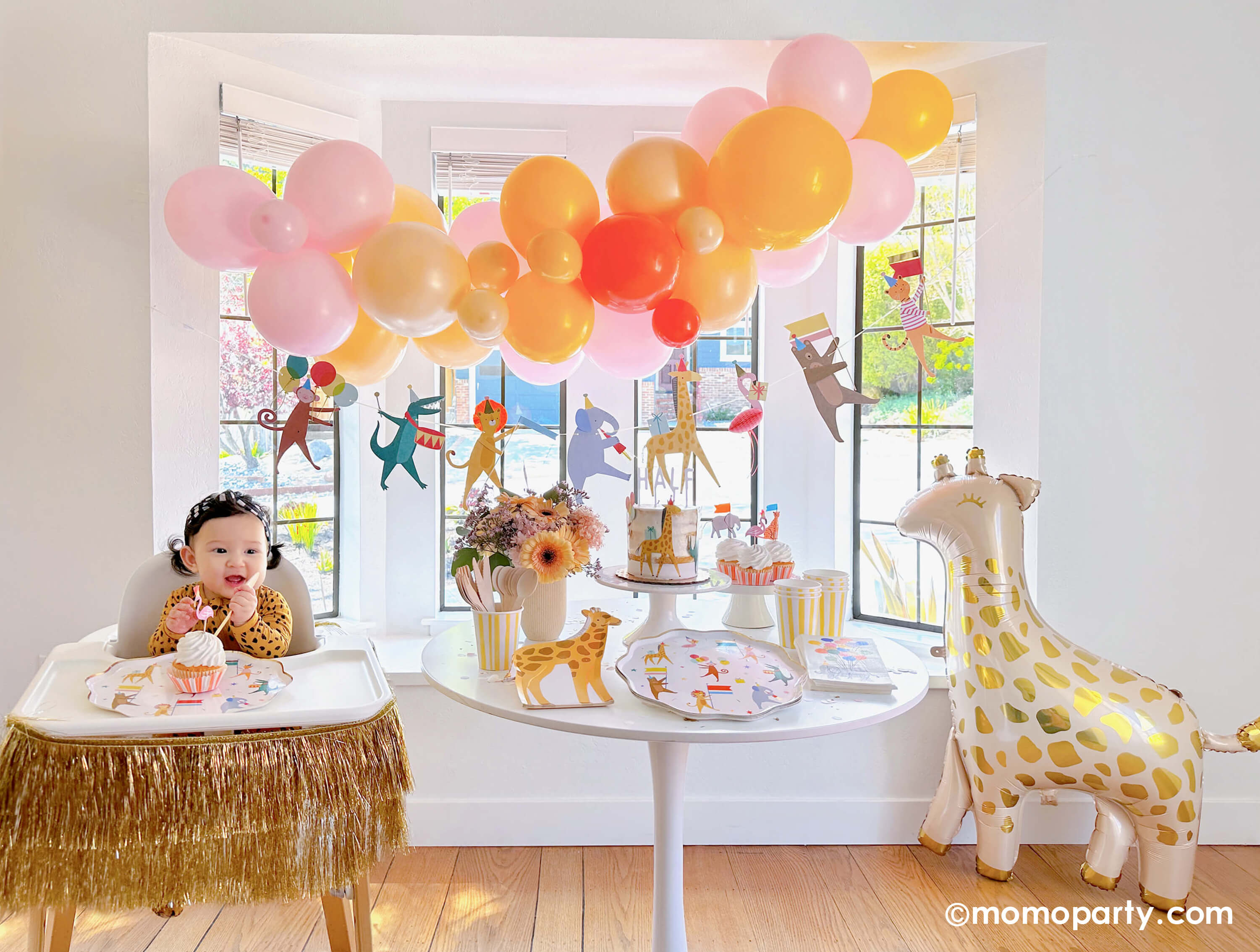 An adorable baby's animal themed 6 months party featuring Momopartys's animal parade birthday garland featuring a lion, an elephant, an alligator, a giraffe, along with a festive balloon garland in a warm color tone hanging above. On the dessert table, there are Meri Meri's animal parade dinner plates, giraffe shaped plates and cupcakes dressed with the safari animal toppers, next to the table there's a giraffe shaped foil balloon. One the left a highchair is decorated with a gold tinsel garland.