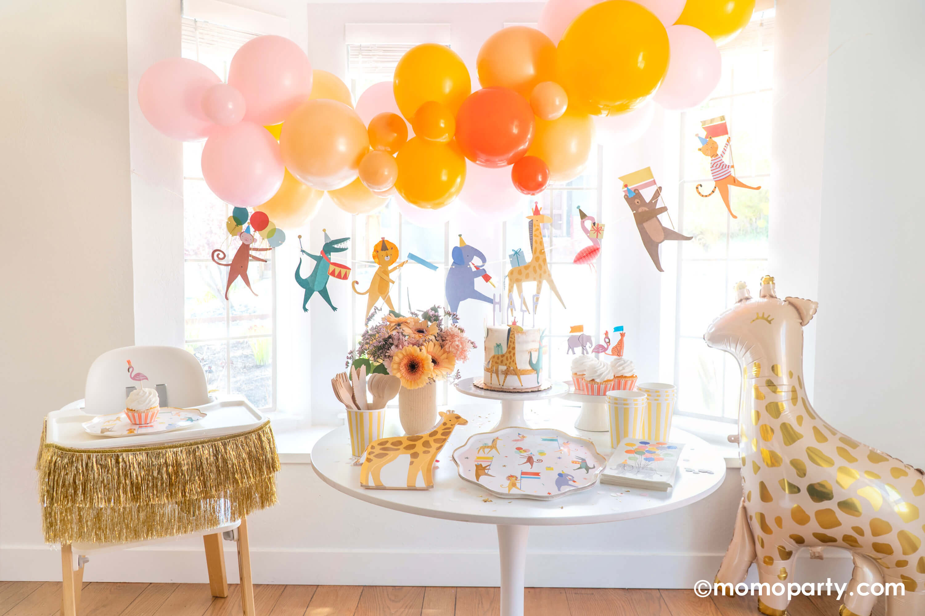 Adorable baby's animal themed party featuring Momopartys's animal parade birthday garland featuring a lion, an elephant, an alligator, a giraffe, along with a festive balloon garland in a warm color tone hanging above. On the dessert table, there are Meri Meri's animal parade dinner plates, giraffe shaped plates and cupcakes dressed with the safari animal toppers, next to the table there's a giraffe shaped foil balloon. One the left a highchair is decorated with a gold tinsel garland.