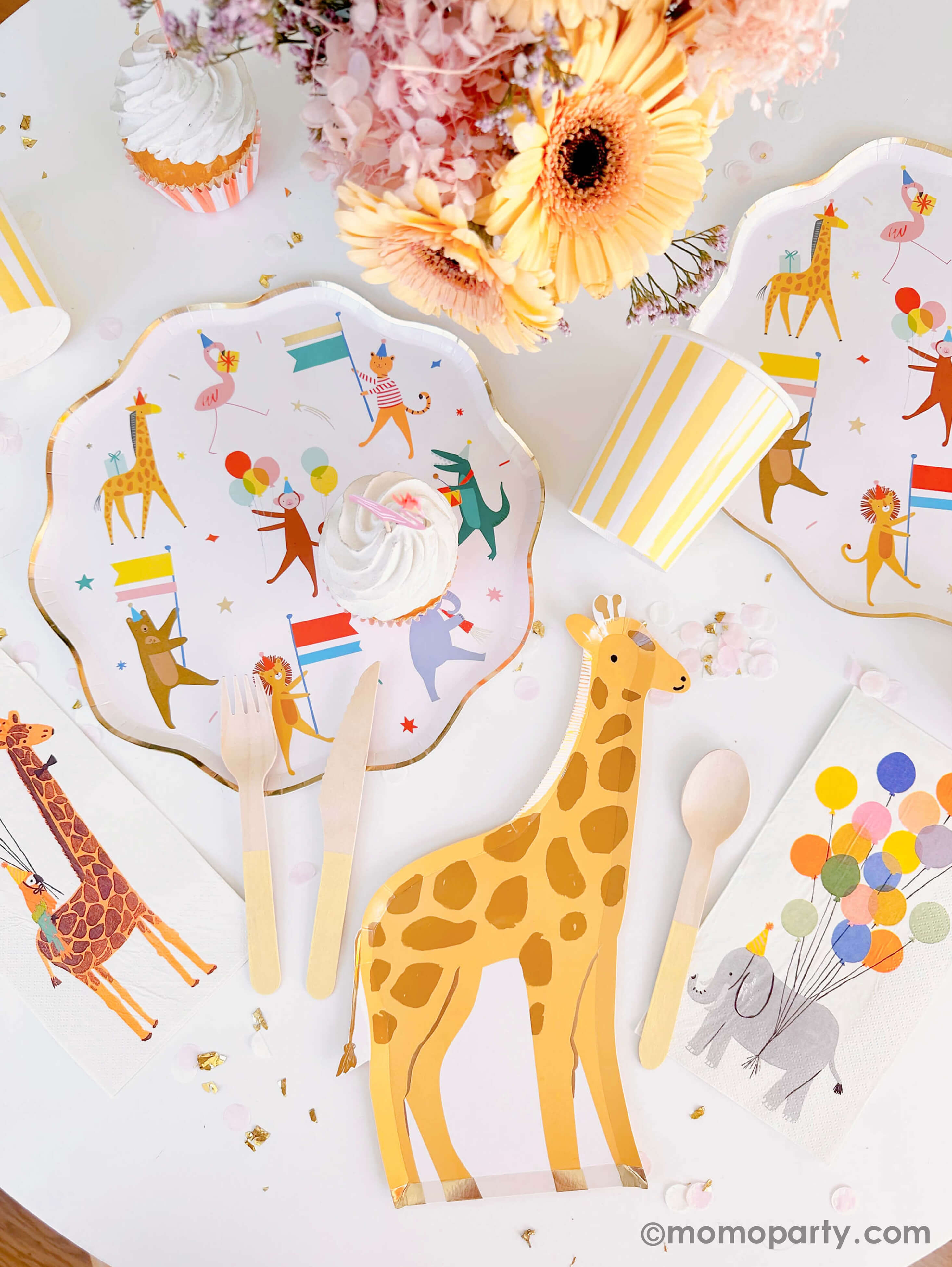 A kid's animal themed tablescape featuring Momopartys's Animal Parade Dinner plate with gold scallop edge and cute animal illustration designs, a giraffe shaped plate for cakes by Meri Meri, and Rifle Paper Co's party animal guest napkins with a cute illustration of an elephant with a party hat and bunch of festive balloons, along with light yellow striped cups and wooden utensil, it makes a great party inspiration for kid's animal, safari, carnival themed birthday party.