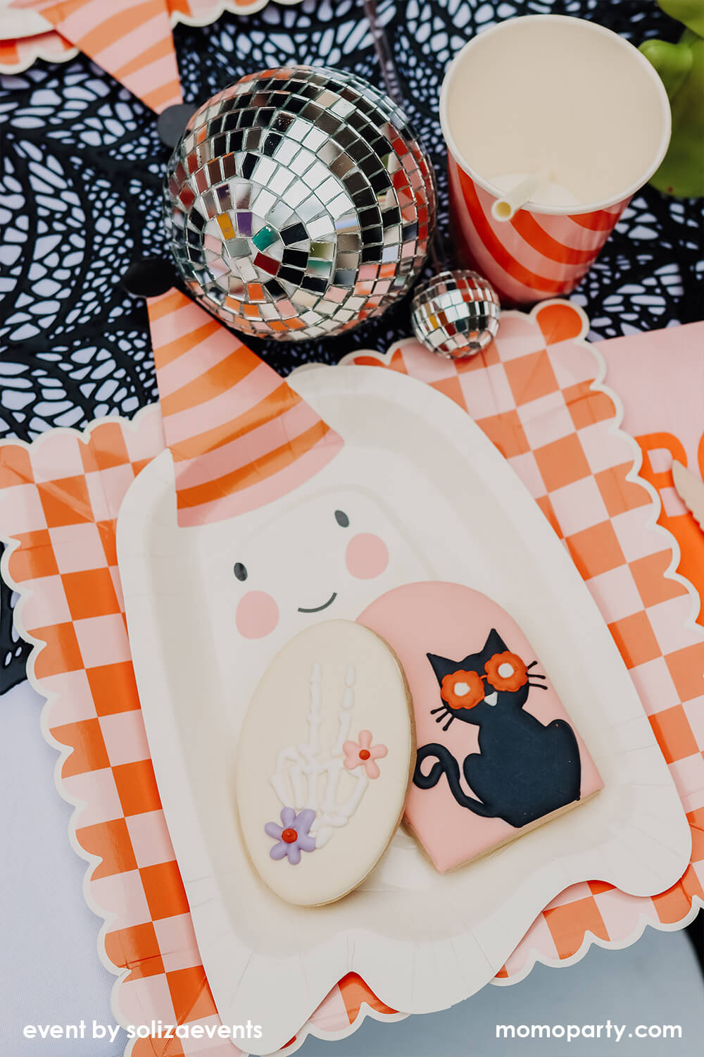 Details of Momopartys Groovy Halloween Themed Party Table, featuring a cookie with black cat and skeleton hand design on the My Mind's Eye Ghoul Gang Ghost Shaped Paper Plates, laid over Ghoul Gang Checkered Paper Plates, Iridescent Party Straws inside the Meri Meri Pink & Orange Stripy Cups, Shining Disco balls, They all on the black spider web placemats. This is such a modern fun Halloween theme and Halloween party for kids.