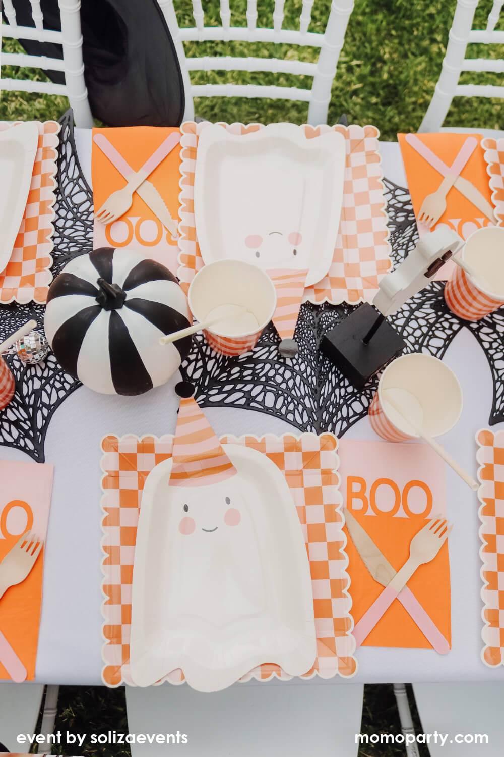 Top view of Momopartys Groovy Halloween Themed Party Table, decorated with 6 sets of My Mind's Eye Ghoul Gang Ghost Shaped Paper Plates, over Ghoul Gang Checkered Paper Plates, Happy Haunting Boo Guest Towels, Iridescent Party Straws inside the Meri Meri Pink & Orange Stripy Cups, They all on the black spider web placemats, and there is a black and white striped pumpkin, a cute ghost Ghost Cutout with a Boo base as the centerpiece. This is such a modern fun Halloween theme and Halloween party for kids.
