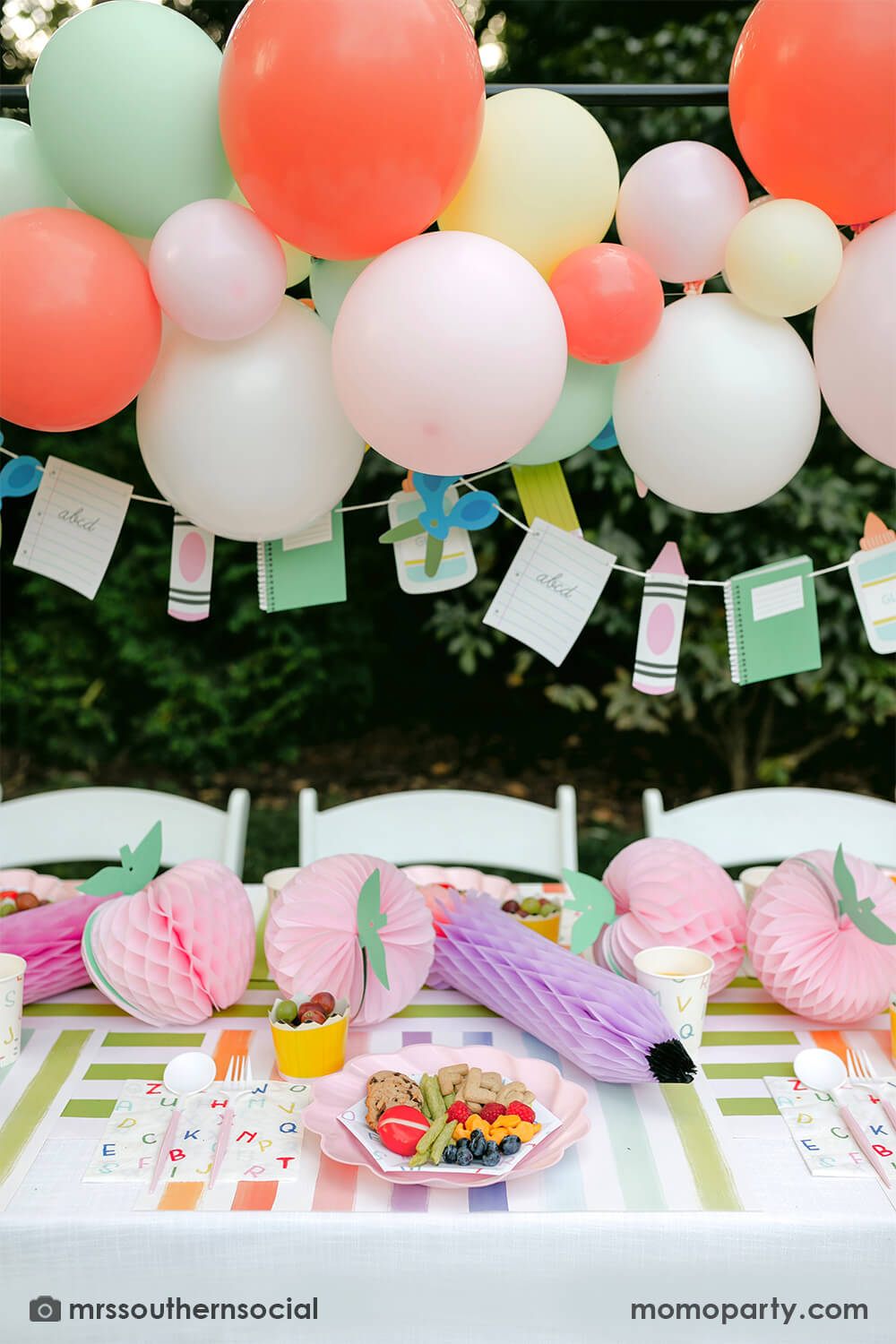 A backyard pastel themed back to school party table set up and decoration featuring Momopartys's back to school themed party goods including back to school party garland, pink apple honeycombs and rainbow pastel pencil honeycombs as the centerpiece, ABC alphabet plates, party cups and napkins. With a pastel themed balloon garland hung above the table, it makes an adorable celebration for kid's first day of school.
