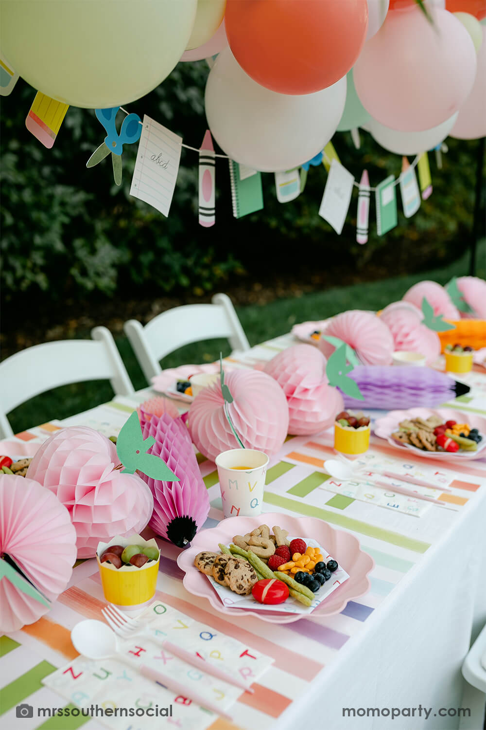 A backyard pastel themed back to school party table set up and decoration featuring Momopartys's back to school themed party goods including back to school party garland, pink apple honeycombs and rainbow pastel pencil honeycombs as the centerpiece, ABC alphabet plates, party cups and napkins. With a pastel themed balloon garland hung above the table, it makes an adorable celebration for kid's first day of school.