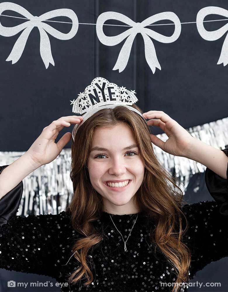 A young lady in a black sequin top wearing Momopartys's NYE silver glittered crown headband by My Mind's Eye. In her back hung a silver tinsel party banner and a silver glittered bow party garland, making this a modern and chic New Year's Eve countdown party decoration inspiration for this festive Holiday season.