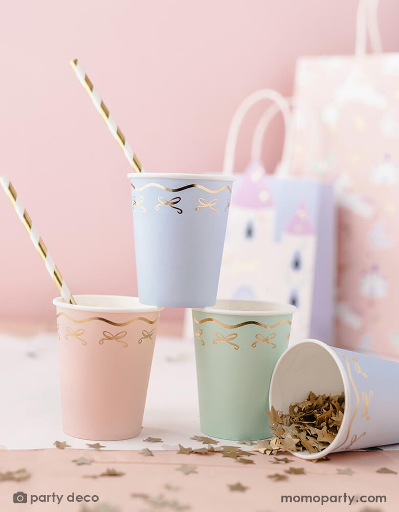 A pink party table featuring Momopartys's 7.4 oz pastel bow party cups in 3 different pastel colors with gold striped straws by Party Deco. Around the cups, there's some gold star confetti spread across the table. In the back there are some girl's party bags.