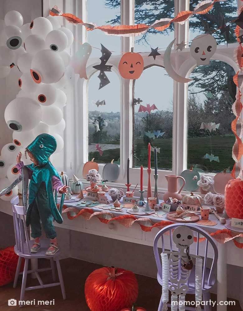 A not-so-spooky Halloween party decoration set up by Meri Meri featuring Momopartys's pastel Halloween party supplies including an eyeball balloon garland, pastel Halloween stitched streamer and pastel Halloween large garland hung on the wall. On the table was set with various pastel Halloween themed tableware including pumpkin shaped plates, napkins and party cups. In the front two kids dressed up in costume having fun at the party.