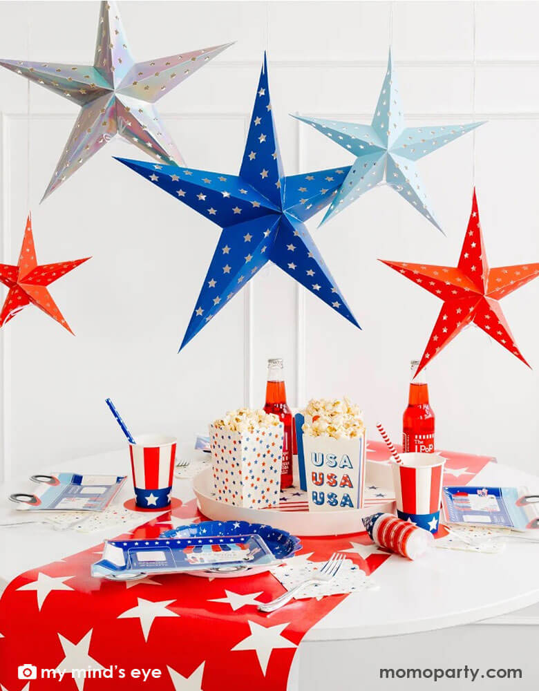 A festive Fourth of July party table decoration featuring Momopartys's Sparklers and Rockets Decorative Hanging Stars by My Mind's Eye hanging above the party table filled with stars and stripes themed patriotic party supplies including a red star pattern table runner, firework truck shaped plates, uncle sam hat party cups, rocket pop treat poppers and star patterned treat boxes with popcorn. All makes a great inspo for an unforgettable 4th of July celebration!