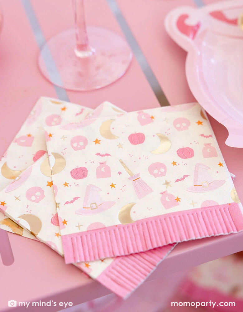 A close up shot of a pink table featuring Momopartys's pink witch Halloween party supplies including the pink Hocus Pocus fringe small napkins by My Mind's Eye. Featuring a fun "Hocus Pocus" pattern including witch themed designs like potion, witch's hats, skulls, bats, pumpkins, crescent moons, stars and fringe detailing, along with the pink cauldron shaped plates on the table, they're perfect for a chic pink witch themed Halloween celebration.