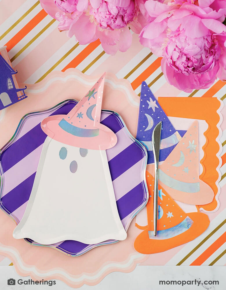 A festive pink Halloween party table featuring Momopartys's Bewitching ghost shaped plates, purple striped dinner plates, bewitching witch shaped hat napkins in three different colors of purple, orange and peach laid on a pink, orange and gold striped table runner. There are some beautiful peony flower arrangement in the middle of the table as the centerpiece, making this an adorable inspo for a chic and cute Halloween table for this spooky season!