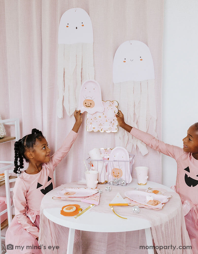 Two school aged girls in pink shirts that have Jack-o-lantern design on them and pink tutu sitting at a party table holding Momopartys's ghost shaped party plates. On the party table there are multiple pink Halloween party tableware spread out and the side is adorn with the pink ghost felt banner. Under the table there are some big disco balls and in the back hung some ghost paper decorations with streamer - a sweet and cute decoration for a Halloween celebration that is kid's friendly.