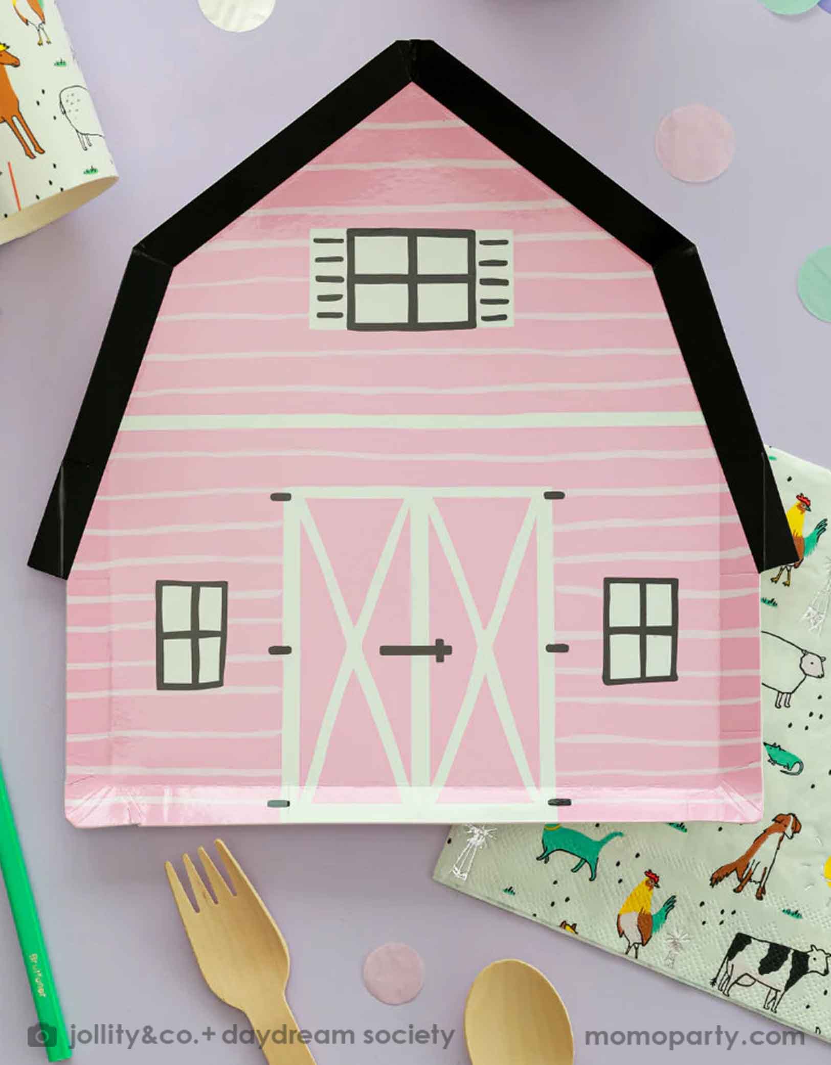 A kid's farm themed birthday party tablescape featuring Momopartys's pink on the farm barnhouse shaped plate and farm motif large napkins and party cups with farm animals including cows, chickens, dogs, pigs etc, with pink confetti and wooden cutlery around.