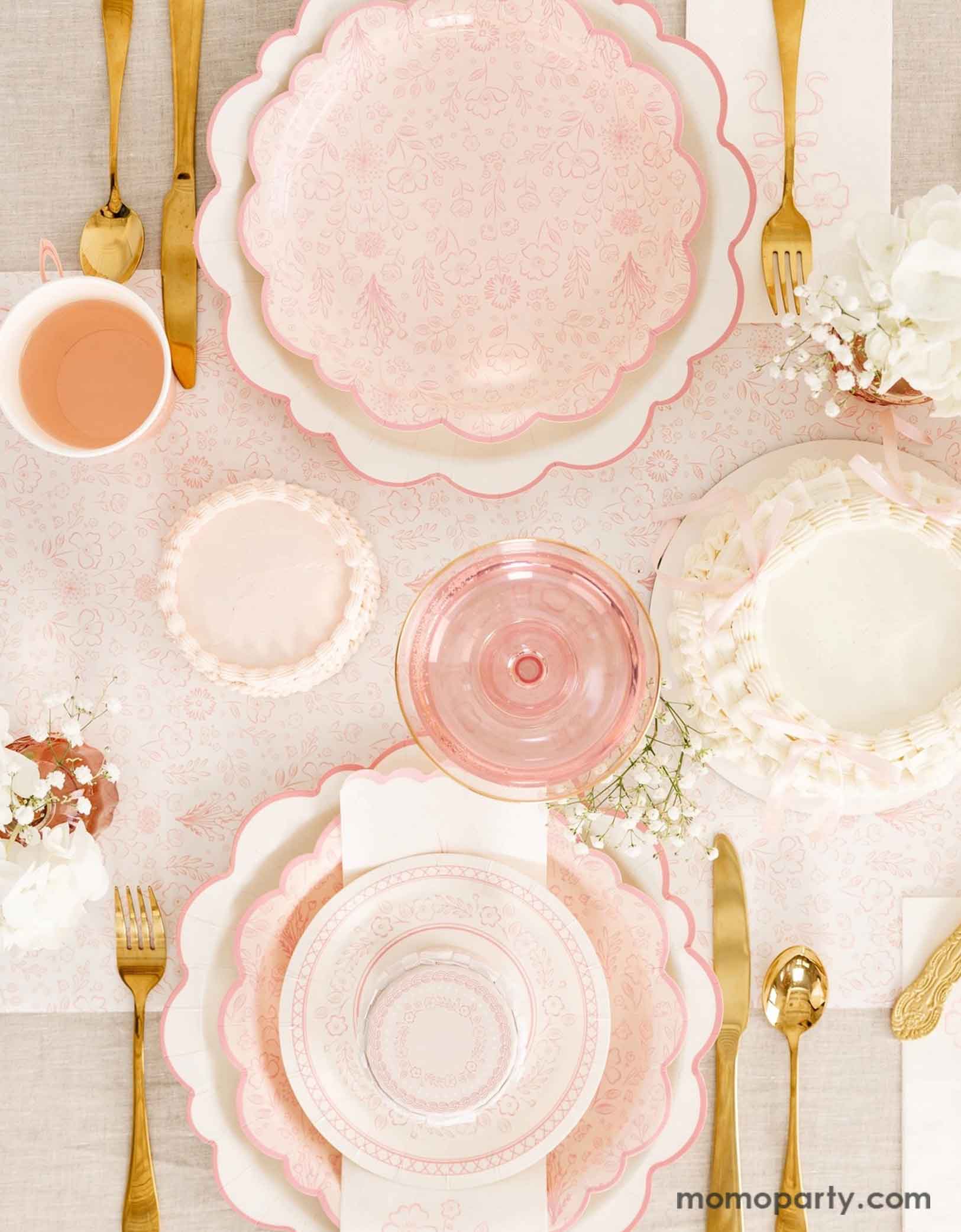 An elegant tablescape featuring Momopartys's Pink Pembroke tableware collection including the 12" Pembroke Pink Edge Scallop Plates paired, 9" Pembroke Pink floral Plates, 7" Pembroke Pink Plates, Pembroke Pink napkins and party cups. Underneath the plates is the Pembroke Pink floral paper table runner. With the gold foil utensil set, a pretty floral arrangement and pastel pink butter cakes and glasses of rose champagne, this table setting is perfect for an Easter celebration or Mother's Day party.