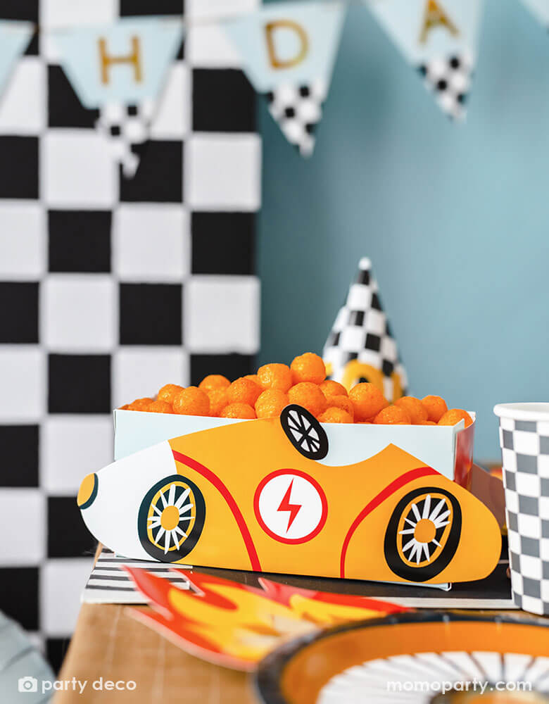 A modern kid's race car themed party table features Momopartys's race car snack box in yellow, filled with cheese ball snacks. On the table there are checkered pattern party cups, party hats and wheel shaped plates by Party Deco on a race track table runner. In the back there's a checkered backdrop decorated with a trophy "Happy Birthday" party banner in light blue, making this a perfect inspo for kid's race car themed parties.