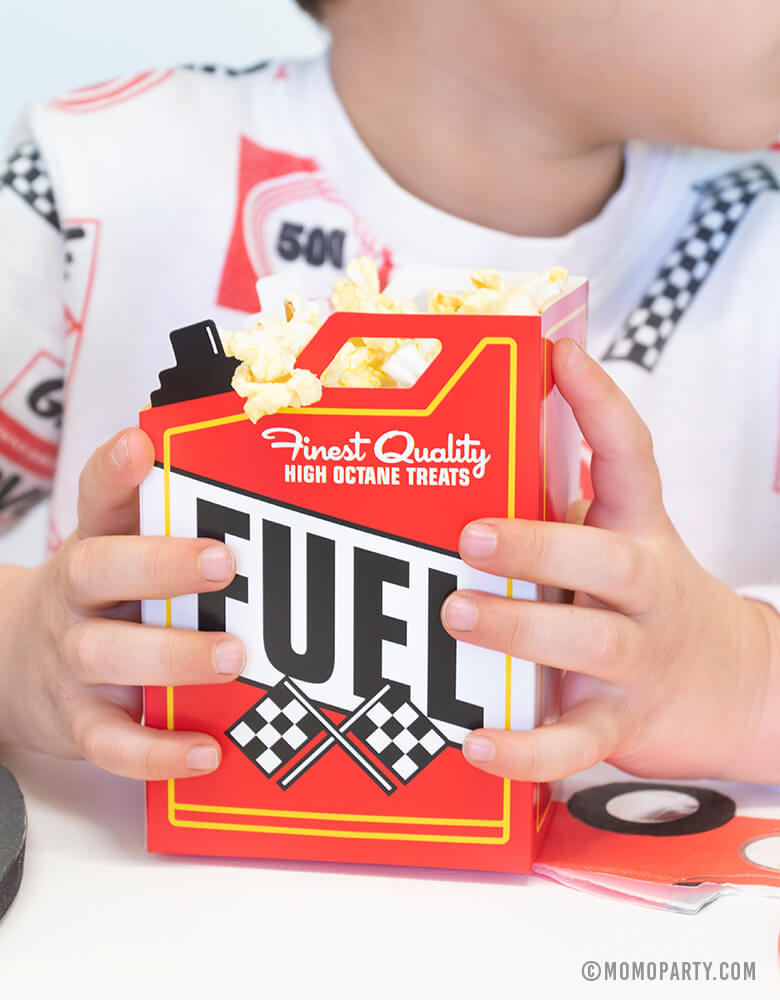 A boy dressed in a race car themed sweat shirt holding Momopartys's fuel snack box filled with popcorn in a race car themed party.