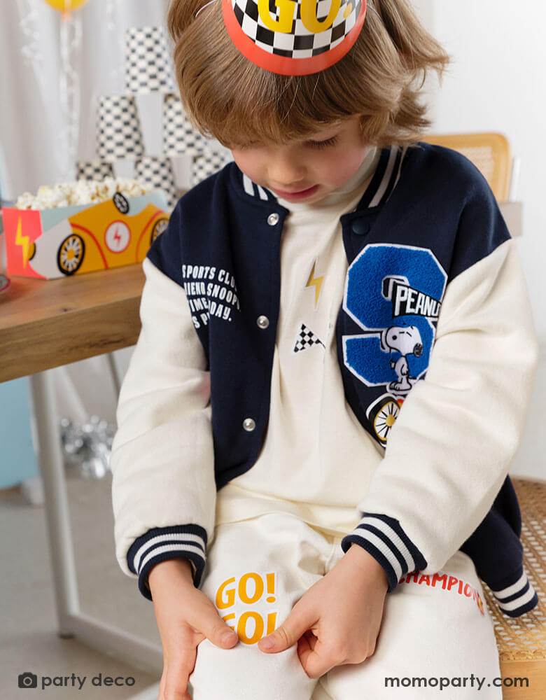 A little boy wearing a checkered flagged party hat with GO! on it in a race car themed birthday party. In his back is a party table filled with race car themed treat boxes with popcorn in them and some checkered patterned party cups stacked. He's putting Momopartys's race car sticker by Party Deco on his clothes - a perfect party activity for boy's race car themed birthday celebrations.