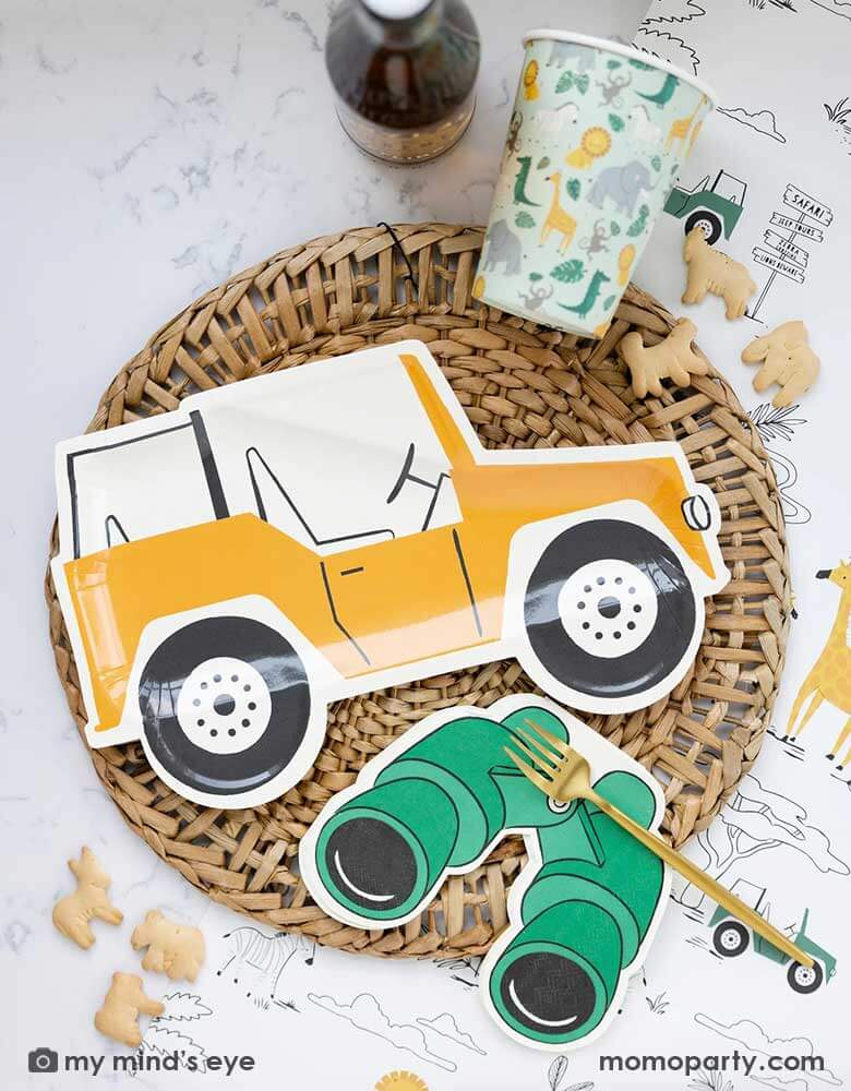 A safari adventure themed party table featuring Momopartys's mustard Jeep shaped dinner plate laid on top of a round bamboo weaved round tablemat which is placed on My Mind's Eye's safari adventure paper table runner featuring safari animals and safari landscape. Around the plate are a mint party cups with lots of safari animals and Momopartys's green binoculars shaped dinner napkins. With animal crackers on the table, makes this tablescape a great inspiration for kid's safari themed party.