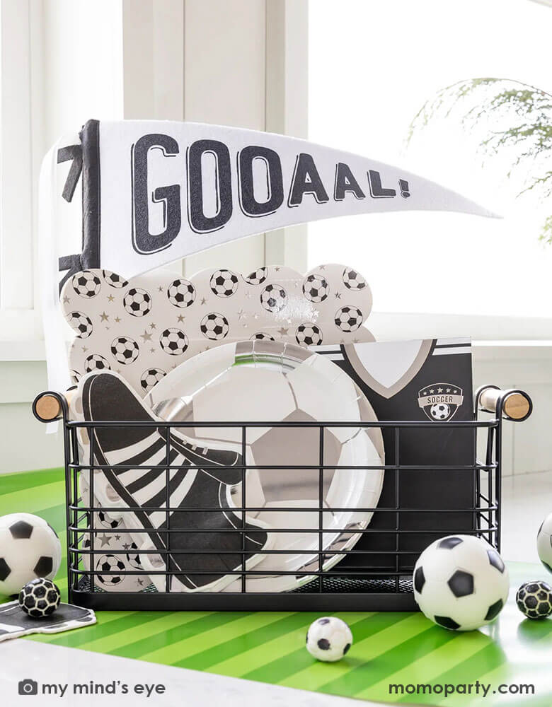 A festive occasions bin for a fun soccer party featuring Momopartys's silver soccer ball shaped plates, soccer cleats shaped napkins, black soccer jersey shaped treat bags and a black and white GOAL felt party pennant by My Mind's Eye. With a green striped paper runner underneath and some soccer ball shaped candies around the table, this makes a perfect inspo for kid's soccer themed birthday party or championship celebration.