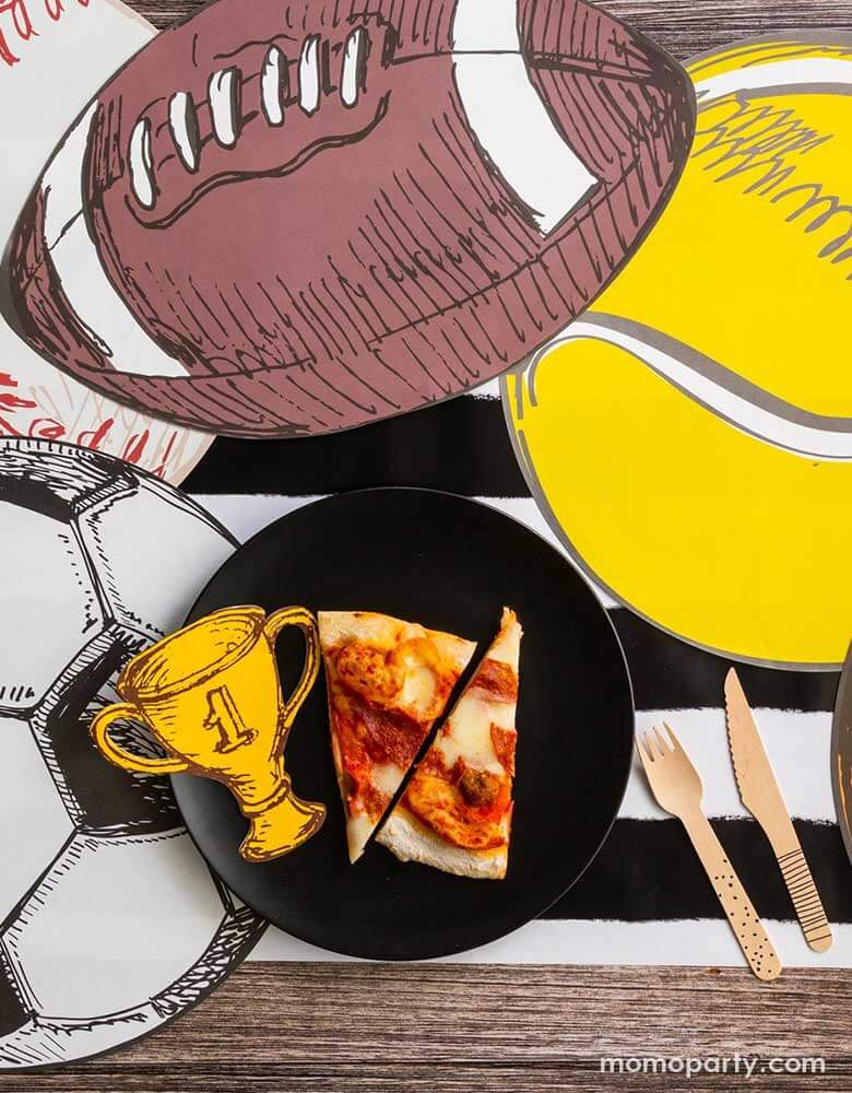 A sports themed party table featuring Momopartys's 7" trophy shaped napkins and a vintage inspired ball shaped placemats including a football placement, a tennis ball shaped placement, a baseball shaped placemat and a soccer ball shaped placemat. On the table there is a plate with a couple of slices of pizzas and some wooden cutleries, all on a black and white striped table runner.