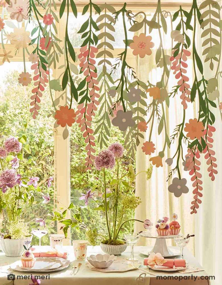 A spring inspired party table set up next to a large window with garden in the back. On the table there are some spring floral cupcakes, party crackers and party tableware, above the table hung Momopartys's floral paper backdrop which features lots of flower and foliage paper pennants, creating a wow effect to this event space.