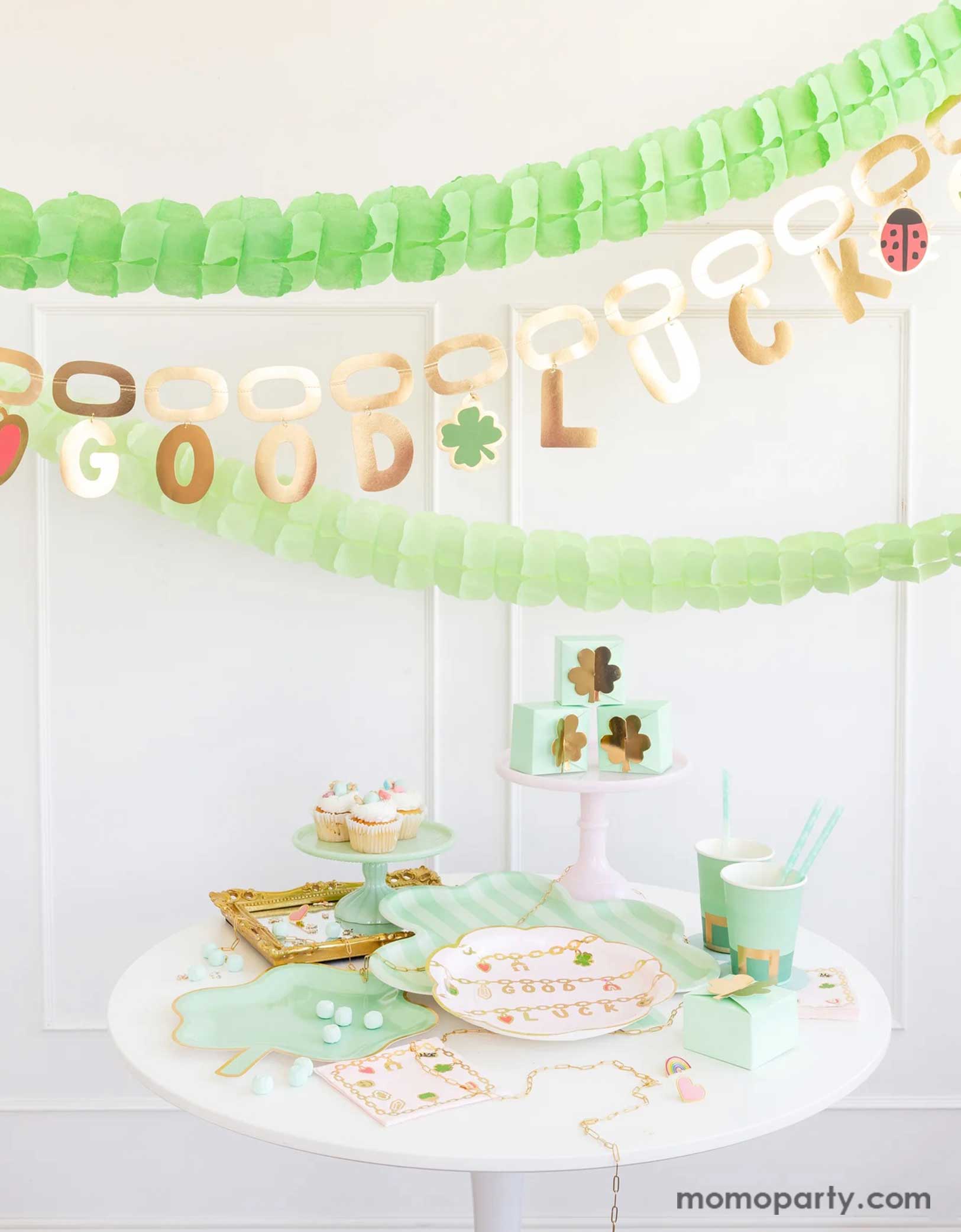 A modern and clean St. Patrick's Day celebration decoration featuring Momopartys's St. Patrick's Day party supplies including shamrock shaped plates, bamboo serving tray, leprechaun shaped party cups, lucky charm scalloped plates and shamrock treat boxes. Above the table hung the good luck charm party banner in gold foil. Along with green tissue honeycomb banner, this creates a charming scene for an adorable St. Patrick's Day celebration for guests of all ages!
