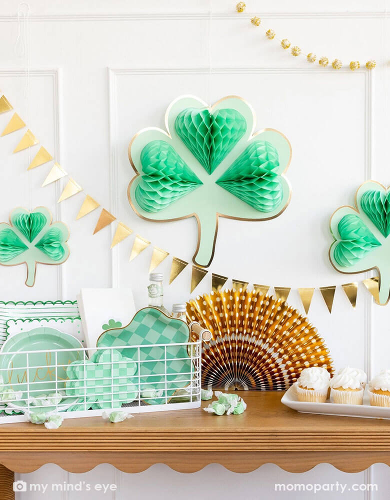 A corner of a room decorated with Momopartys's shamrock honeycomb decorations in various sizes and a gold foil flag banner hung on the wall. On the cabinet counter, there's a gold foil paper party fan, a few cupcakes and a basket filled with Momopartys's St. Patrick's Day party tableware including scalloped paper plates, lucky paper plate set, checkered shamrock shaped napkins and plates next to some green taffy candies, makes this a perfect info for a festive St. Patrick's Day celebration.