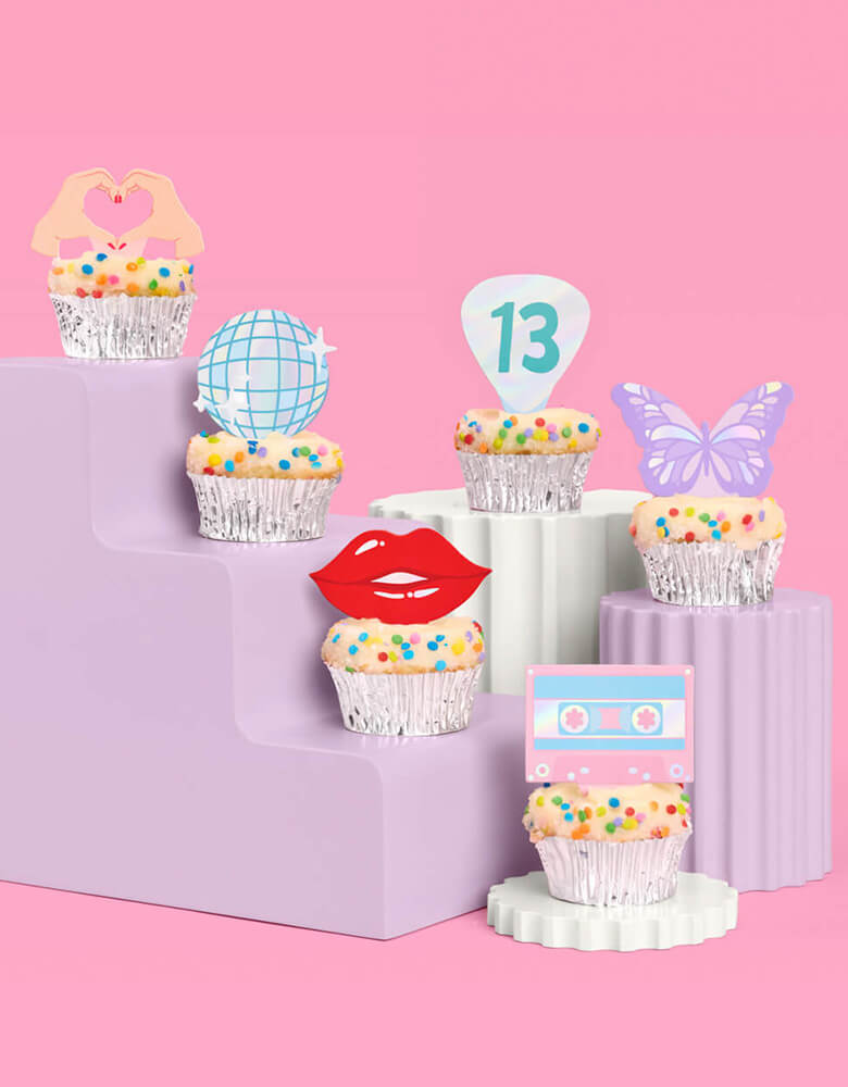 A few birthday cupcakes adorned with Momopartys's Era's Party Cupcake Toppers by Xo, Fetti on a modern lilac cupcake stand. Comes in a set of 24, these Taylor Swift-themed dessert/cupcake toppers are with iconic designs including butterflies, red lips, disco balls, cassettes, guitar picks and heart hand symbols. They're perfect for to celebrate your Swiftie girl's special birthday.