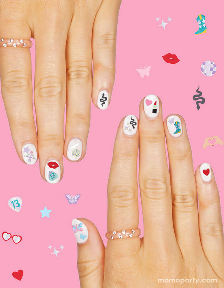 Two hands with white nail polish decorated with Momopartys's Taylor Swift Era's Nail Stickers by Xo, Fetti. Each pack comes with 544 adhesive nail stickers in the Taylor Swift inspired designs including red lipstick, red lips, butterflies, cowgirl boots, snakes, cassette tapes, guitar picks, heart hand symbols, and heart shaped sunglasses. These nail stickers make perfect party activity for your Swiftie girl's special birthday celebration.