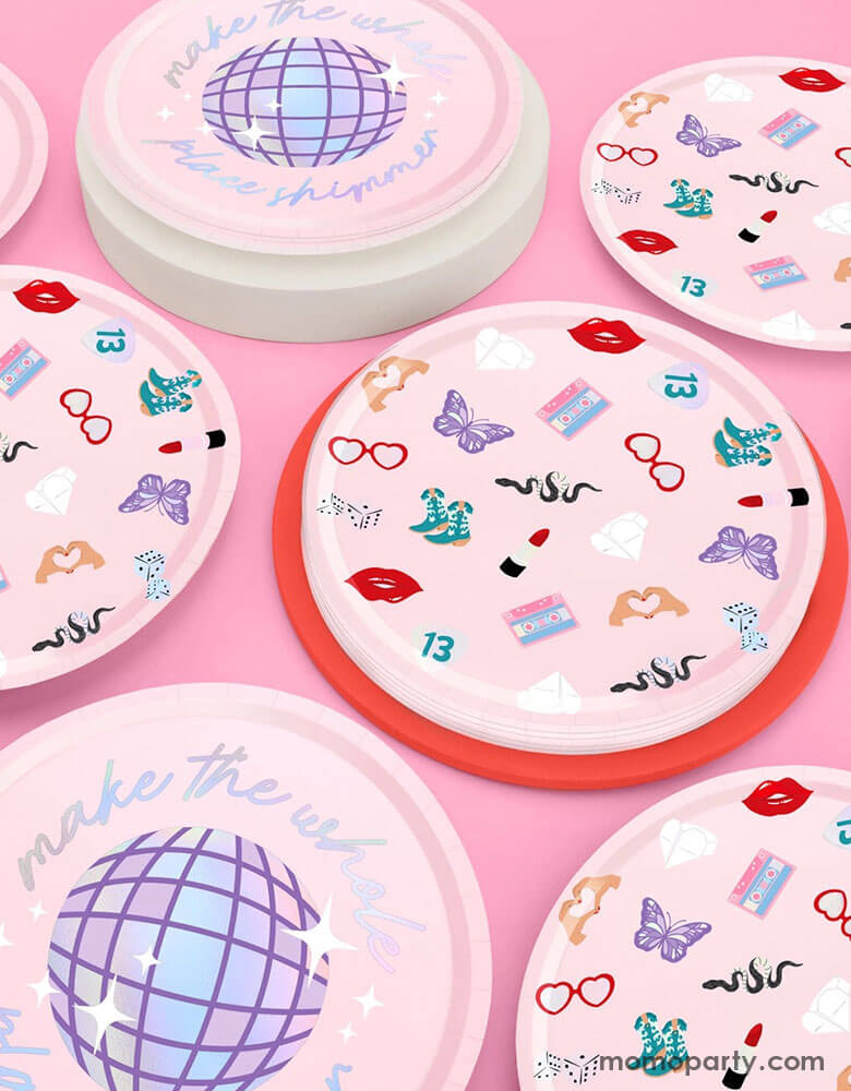 Momo Party's 9" x 9" Taylor Swift Era's Party Paper Plates by Xo, Fetti. Comes in a set of 24 paper plates in two different designs, a disco ball and a Taylor Swift inspired motif featuring red lipstick, red lips, butterflies, cowgirl boots, snakes, cassette tapes, guitar picks, heart hand symbols, and heart shaped sunglasses. They're perfect for the party table celebrating your Swiftie girl's special day.