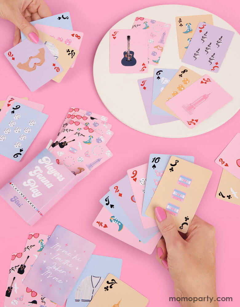 Two ladies in their pink nail polish playing Momopartys's Taylor Swift Era's Tour Party Playing Cards by Xo, Fetti. Each pack has a full deck of 52 playing cards + 2 jokers in light purple, pink, blue and peach cards with Taylor Swift inspired designs featuring red lipstick, red lips, butterflies, cowgirl boots, snakes, guitars, cassette tapes, guitar picks, heart hand symbols, and heart shaped sunglasses.
