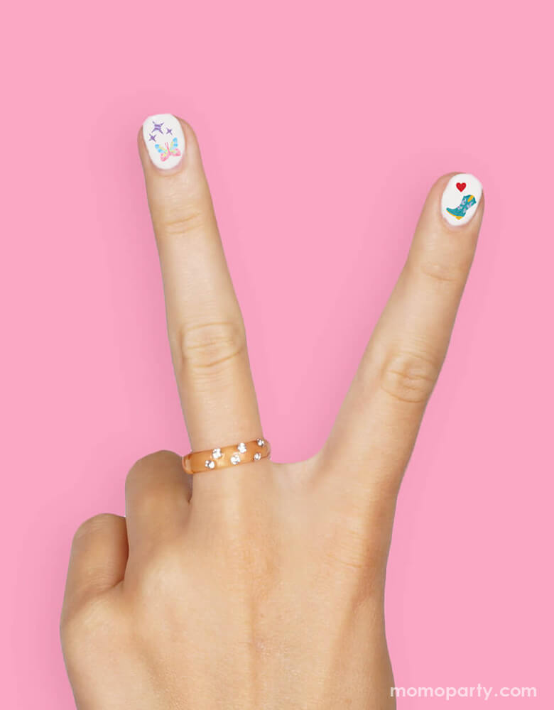 A peace hand gesture with white nail polish decorated with Momopartys's Taylor Swift Era's Nail Stickers by Xo, Fetti. Each pack comes with 544 adhesive nail stickers in the Taylor Swift inspired designs including red lipstick, red lips, butterflies, cowgirl boots, snakes, cassette tapes, guitar picks, heart hand symbols, and heart shaped sunglasses. These nail stickers make perfect party activity for your Swiftie girl's special birthday celebration.